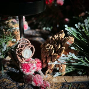 May include: A handcrafted copper wire ring featuring a clear, round gemstone. The ring is intricately woven with decorative swirls. The ring rests on a sprig of red berries and is next to a pinecone and evergreen branches.