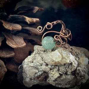 May include: A handcrafted copper wire ring featuring an aquamarine-colored, round gemstone. The ring's design incorporates intricate wire wrapping and swirling patterns. The ring is displayed on a textured, light-colored rock surface, with a dark background.