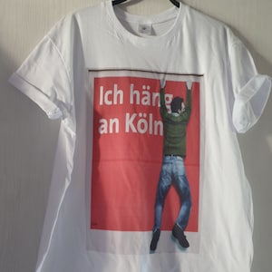 May include: White t-shirt with a red and white graphic print of a person hanging from a wall with the text "Ich häng an Köln".