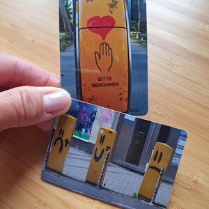 May include: Two yellow street signs with different designs. The first sign has a red heart and a hand with the text "Bitte Berühren" which translates to "Please Touch". The second sign has three yellow poles with different smiley faces.