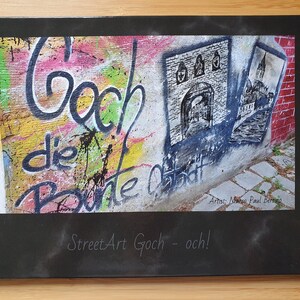 Könnte beinhalten: Ein Schwarzweißfoto einer Backsteinmauer mit bunten Graffitis. Die Graffitis beinhalten die Worte "Goch die Bunte Stadt" und eine Schwarzweißzeichnung eines Gebäudes. Das Foto ist in einem schwarzen Rahmen mit dem Text "Street Art Goch - och!" und "Artist: Marco Paul Bertina" unten gerahmt.