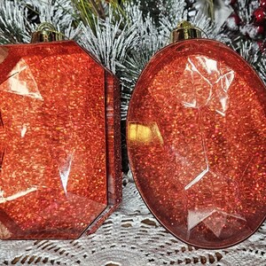 May include: Two red glitter Christmas ornaments shaped like large, faceted gemstones. The ornaments are hanging from a snowy evergreen tree.