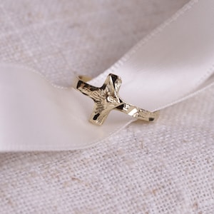 May include: A gold-colored cross ring rests on a white satin ribbon, set against a textured, off-white fabric background. The ring's cross design is detailed, and the ribbon is gently curved.