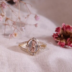 May include: A gold ring featuring an oval-shaped centerpiece with a raised rose gold figure of the Virgin Mary set against a sunburst-patterned silver background, surrounded by small clear stones on the band and the oval frame.