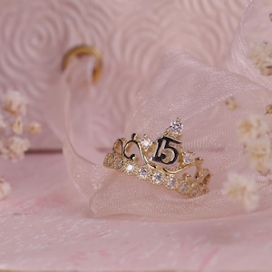 May include: A gold-colored crown ring with the number 15 in black, adorned with clear gemstones. The ring sits on a sheer, light pink fabric, with small white flowers and a blurred background.