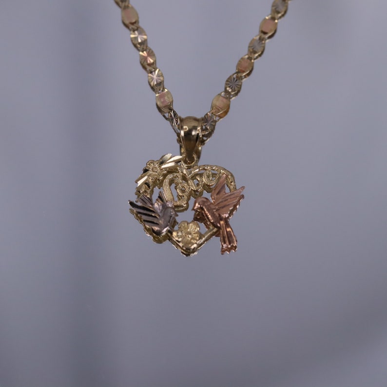 May include: A gold heart-shaped pendant necklace with two birds, one in gold and one in rose gold, perched on a branch with leaves. The chain is a gold, diamond-cut, rope chain.