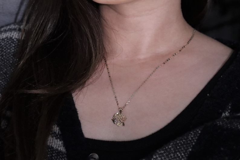 May include: A gold chain necklace with a delicate, intricate pendant hangs on a person's neck. The necklace is worn against a black shirt with a plaid pattern.