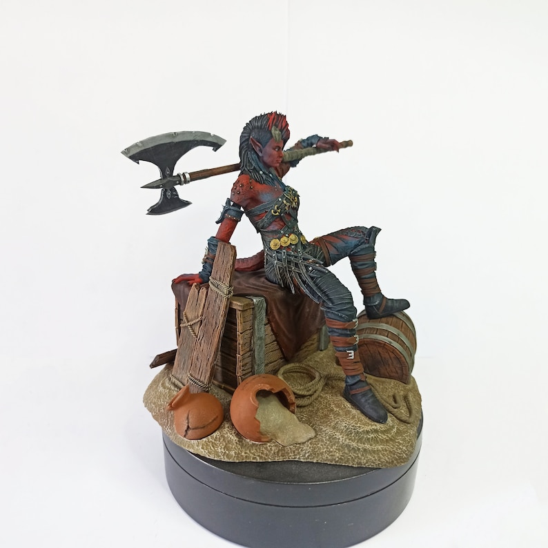 May include: A detailed figurine of a female warrior with red skin, horns, and blue and black armor, holding a large axe. She is seated on a wooden crate with a barrel and broken pottery nearby. The base is a sandy terrain.