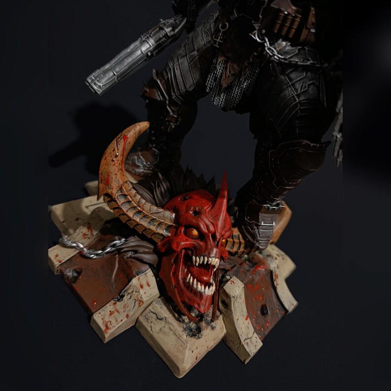 May include: A detailed action figure of a warrior standing on a base with a demon skull. The warrior is wearing dark armour and holding a gun. The demon skull is red with sharp teeth and horns.