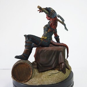 May include: A detailed figurine of a red-skinned character with horns and a tail, dressed in dark armor, seated on a stone block. The figure holds a pickaxe and is posed in a dynamic, action-oriented stance. A wooden barrel is at the base.