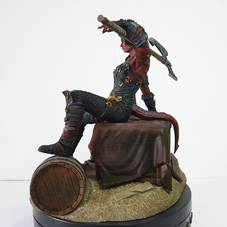 May include: A detailed figurine of a red-skinned character with horns and a tail, dressed in dark armor, seated on a stone block. The figure holds a pickaxe and is posed in a dynamic, action-oriented stance. A wooden barrel is at the base.