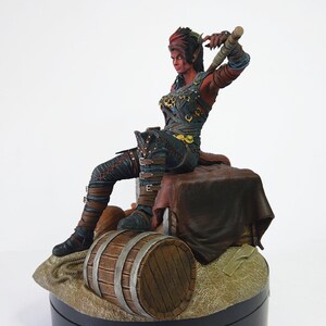 May include: A detailed figurine of a female warrior with red skin, wearing dark armor and seated on a wooden barrel. The figure holds a weapon and is posed dynamically, with intricate details in the clothing and facial features.