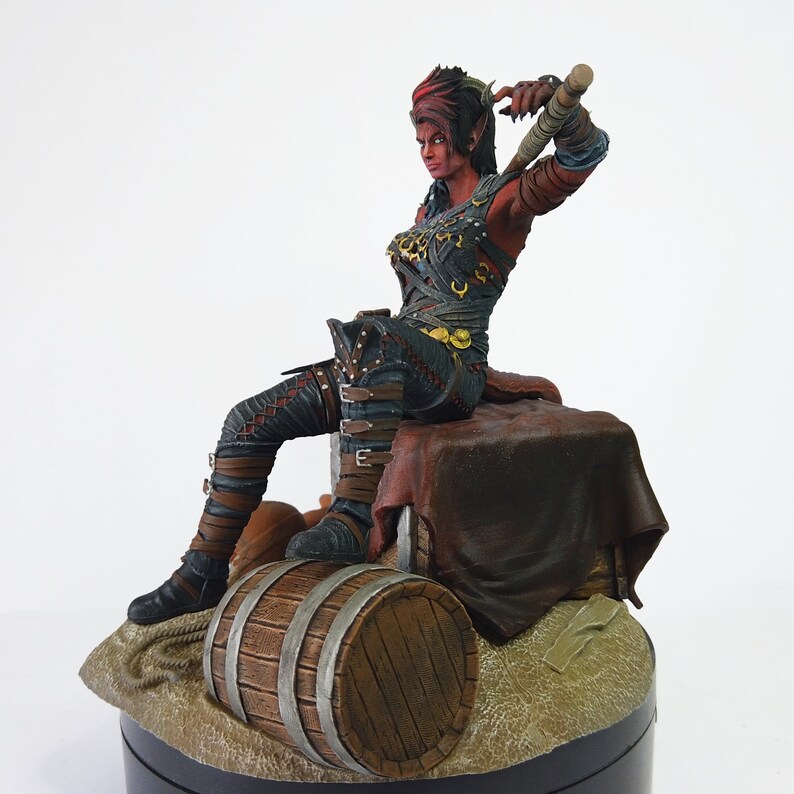 May include: A detailed figurine of a female warrior with red skin, wearing dark armor and seated on a wooden barrel. The figure holds a weapon and is posed dynamically, with intricate details in the clothing and facial features.