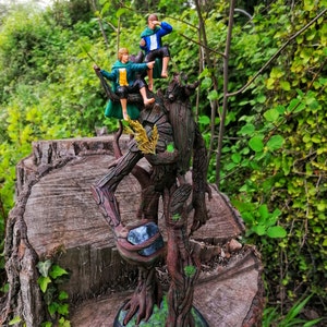 May include: A tree-like figure with a green, brown, and gray paint scheme stands on a tree stump. Two figures are perched on the tree-like figure, one in green and one in blue. The figures are wearing clothing and have painted faces.