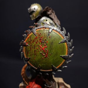 May include: An action figure with a large, circular saw blade shield. The shield is green and silver, with red paint splatters and a symbol in the centre. The figure is wearing a helmet and armour.