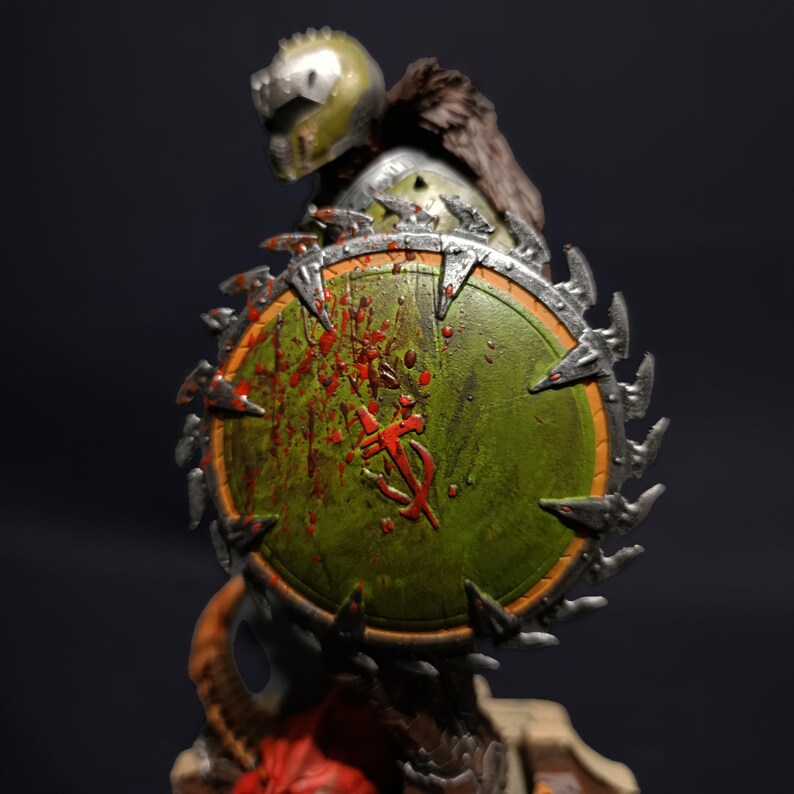 May include: An action figure with a large, circular saw blade shield. The shield is green and silver, with red paint splatters and a symbol in the centre. The figure is wearing a helmet and armour.