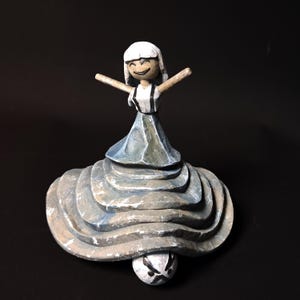 May include: A handcrafted wooden figurine of a smiling person with arms outstretched. The figure wears a white top with black suspenders and a blue skirt, atop a layered, textured base. The base is gray and white, and the bottom has a face.