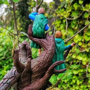 May include: Two miniature figures of people wearing green and blue clothing are perched on a tree branch. The figures are facing away from the viewer and appear to be looking at something in the distance.