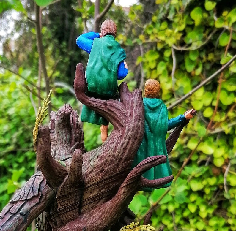 May include: Two miniature figures of people wearing green and blue clothing are perched on a tree branch. The figures are facing away from the viewer and appear to be looking at something in the distance.