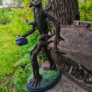 May include: A detailed, hand-painted figurine of a tree creature with a rough, bark-like texture. The creature is holding a small, gray rock in its hand. The figurine is standing on a base with green moss and is painted in shades of brown and green.