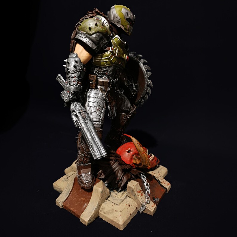 May include: A detailed action figure of a green and silver armoured warrior holding a gun and a chainsaw. The figure is standing on a base with a demon skull.