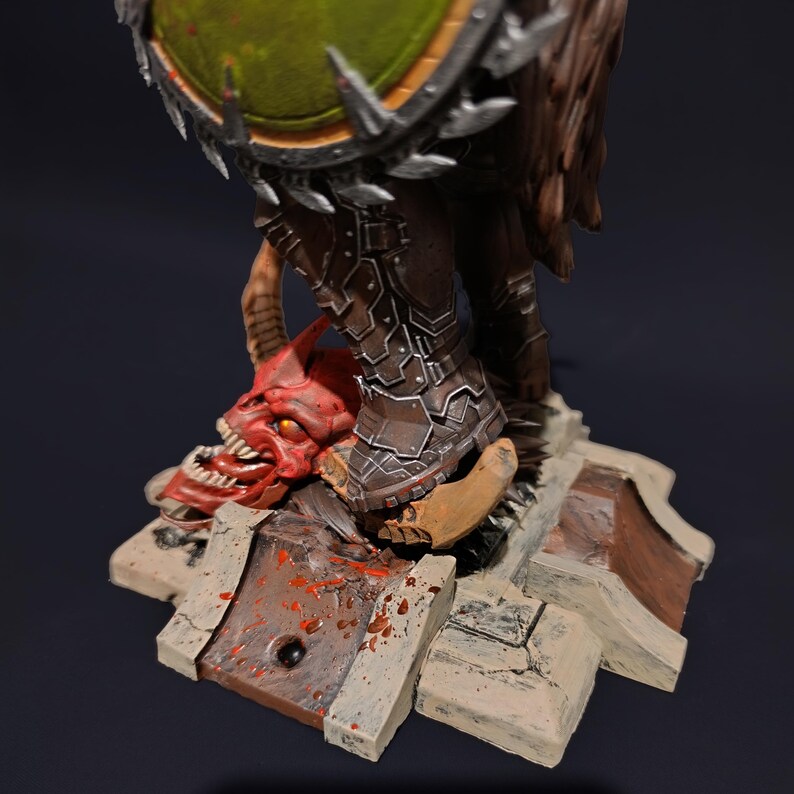 May include: A detailed miniature figure of a warrior standing on a base with a demon's head. The warrior is wearing brown armour with metal accents and is holding a large, green shield with spikes. The base is made of a textured material and is painted in a dark grey colour. The demon's head is red and has sharp teeth.