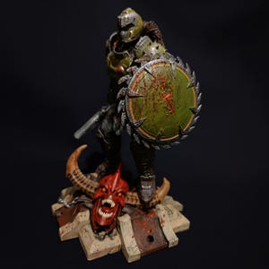 May include: An action figure of a demon slayer, painted in green and grey, holding a chainsaw and a shield with blood splatters. The figure is standing on a base with a red demon head.