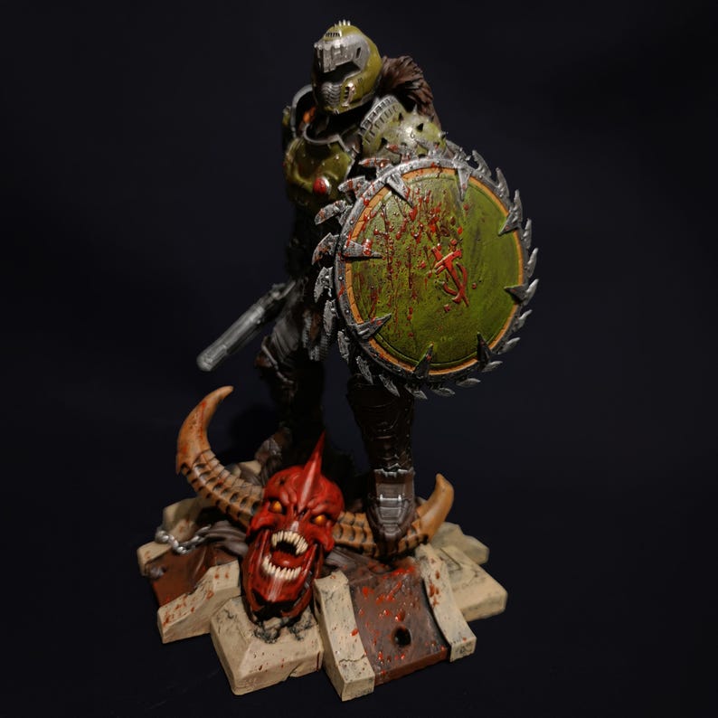 May include: An action figure of a demon slayer, painted in green and grey, holding a chainsaw and a shield with blood splatters. The figure is standing on a base with a red demon head.