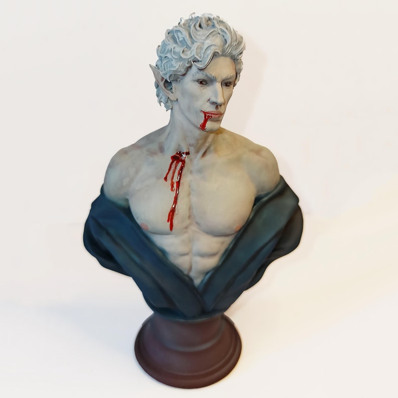 Op de afbeelding: Een witte buste van een mannelijk figuur met puntige oren, een gebeeldhouwde bloedvlek op zijn borst en een blauwe stof die over zijn schouders is gedrapeerd. De buste staat op een bruine basis.