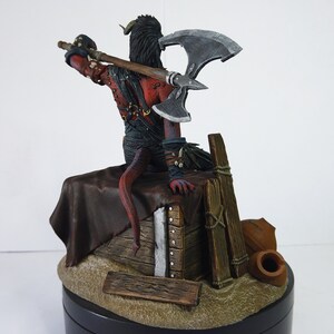 May include: A detailed figurine of a red-skinned demon with horns and a long tail, wielding a large double-bladed axe. The figure is seated on a wooden structure with a brown leather drape, and a small wooden fence. The base is a dark color.