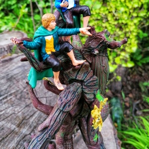 May include: A green and brown tree creature figurine with a small figure of a person in a green and brown outfit sitting on its back. The figure has a yellow vest and is holding a small object in its hand.