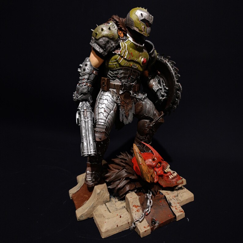 May include: An action figure of a demon warrior, standing on a base with a demon skull. The figure is green and silver, and is holding a gun and a shield.
