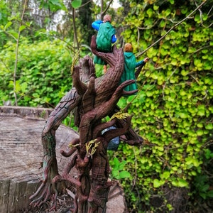 May include: Two figures in green and blue clothing stand on a tree trunk with a gnarled, brown, and textured surface. The figures are standing on a branch that extends out from the trunk. The background is a green leafy bush.