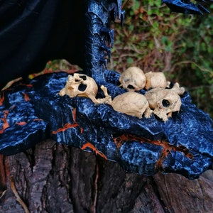 Op de afbeelding: Een zwarte en blauwe rotsbasis met een verzameling kleine, witte schedels. De basis is getextureerd en heeft een ruw, onregelmatig oppervlak.