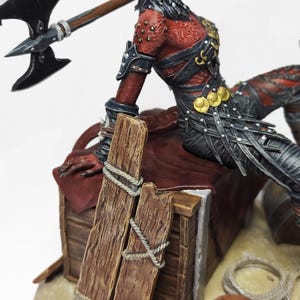 May include: A detailed figurine of a fantasy warrior with red skin and black armor, holding a battle axe. The figure is posed on a wooden structure with rope detailing, suggesting a scene of action or adventure. The axe has a black blade and a wooden handle.