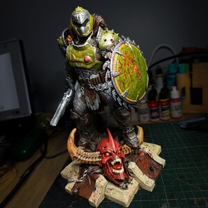 May include: An action figure of a demon, painted in green and silver, wielding a chainsaw and a shield. The figure stands on a base featuring a skull.