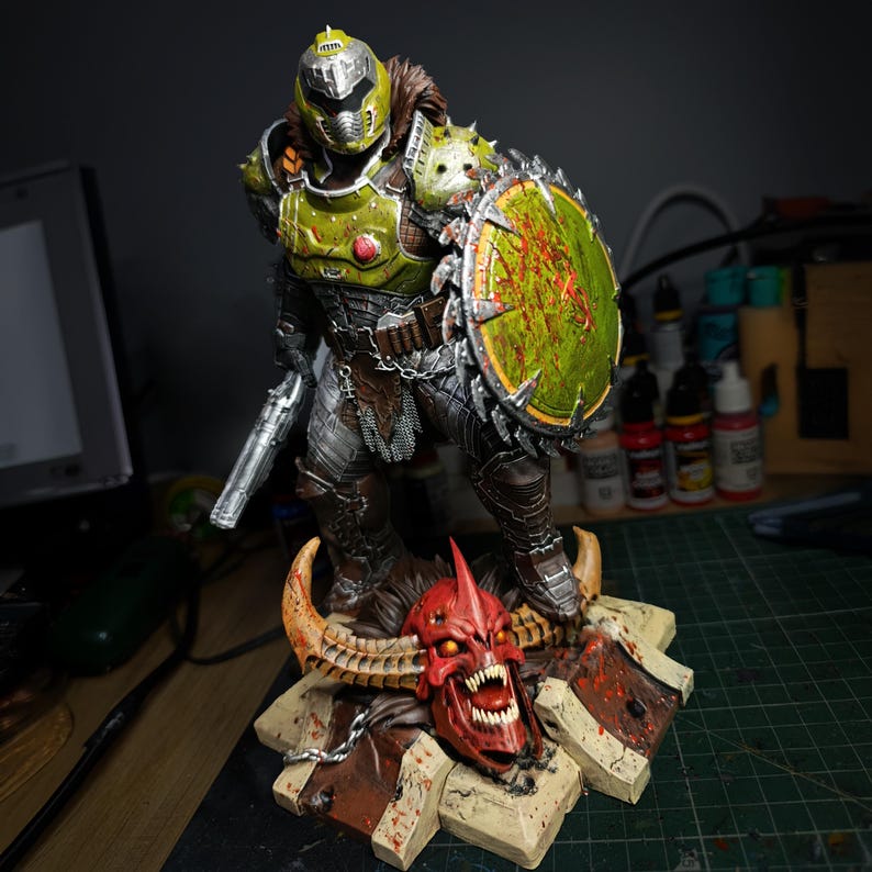 May include: An action figure of a demon, painted in green and silver, wielding a chainsaw and a shield. The figure stands on a base featuring a skull.