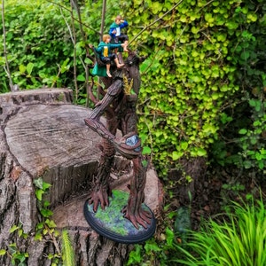 May include: A detailed resin sculpture of a tree with three figures sitting on its branches. The tree is brown and has a rough texture. The figures are wearing green and blue clothing. The sculpture is set against a backdrop of green foliage.