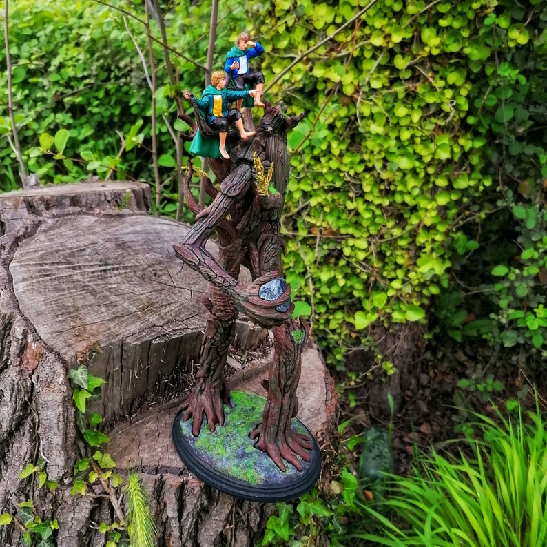 May include: A detailed resin sculpture of a tree with three figures sitting on its branches. The tree is brown and has a rough texture. The figures are wearing green and blue clothing. The sculpture is set against a backdrop of green foliage.