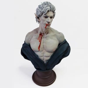 Op de afbeelding: Een bustebeeld van een bleke, mannelijke figuur met puntige oren, die een donkerblauwe robe draagt. De figuur heeft bloed dat van zijn mond en borst druppelt.