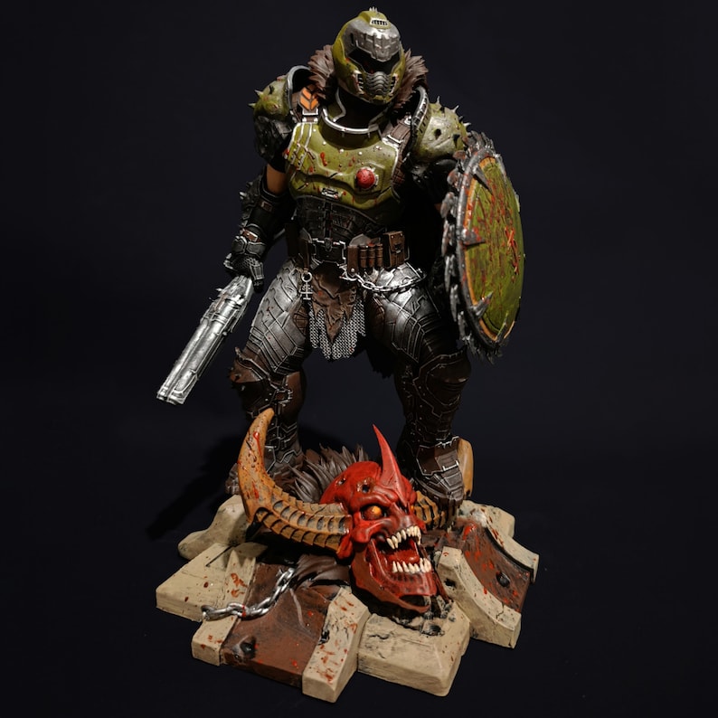 May include: An action figure of a demon warrior, standing on a base with a demon skull. The figure is green and silver, and is holding a gun and a shield.