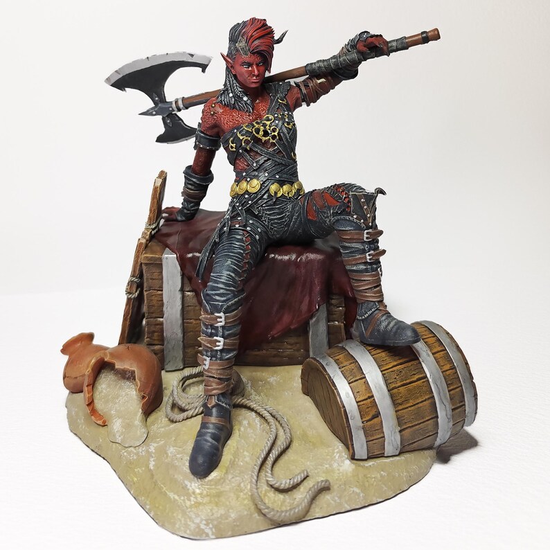 May include: A detailed figurine of a red-skinned warrior with horns, holding an axe. The figure is seated on a small wooden structure with a barrel and saddle, showcasing intricate armor and a dynamic pose. The base is a sandy terrain.