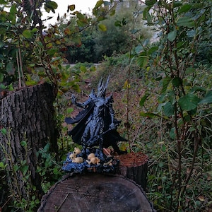 Op de afbeelding: Een zwart figuur van een fantasiepersonage met een cape en zwaard, staand op een boomstronk met een verzameling kleine schedels eromheen. Het figuur bevindt zich in een bosrijke omgeving.