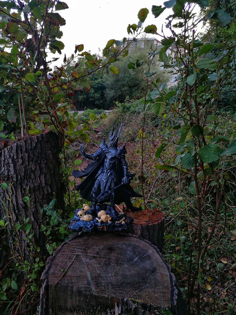 Op de afbeelding: Een zwart figuur van een fantasiepersonage met een cape en zwaard, staand op een boomstronk met een verzameling kleine schedels eromheen. Het figuur bevindt zich in een bosrijke omgeving.