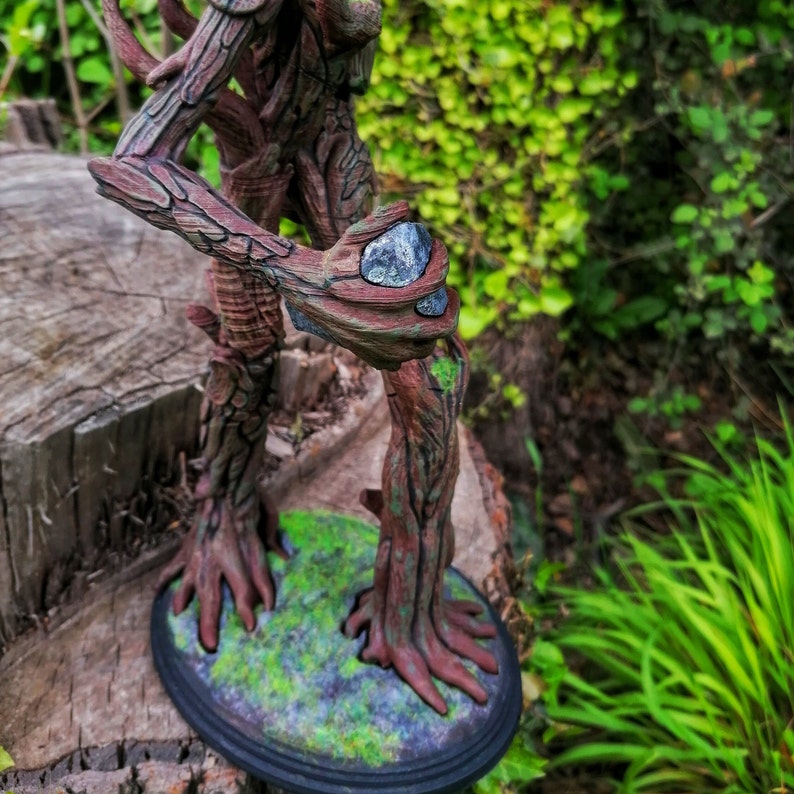 May include: A tree-like figure with brown bark and green moss, standing on a round base. The figure is holding a gray stone in its hand.