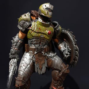 May include: An action figure of a demon warrior, clad in green and silver armour, wielding a chainsaw and a shield. The figure is wearing a helmet with a red visor and has blood splatters on its armour.