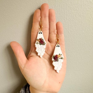 May include: A pair of white ghost-shaped earrings with gold hooks and red, white, and yellow floral accents.