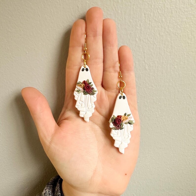 May include: A pair of white ghost-shaped earrings with gold hooks and red, white, and yellow floral accents.