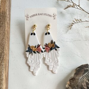 May include: White ceramic ghost earrings with floral details in shades of pink, brown, and black. The earrings are attached to gold-toned hooks. The earrings are displayed on a white card with the text "AMBER RAE STUDIO".