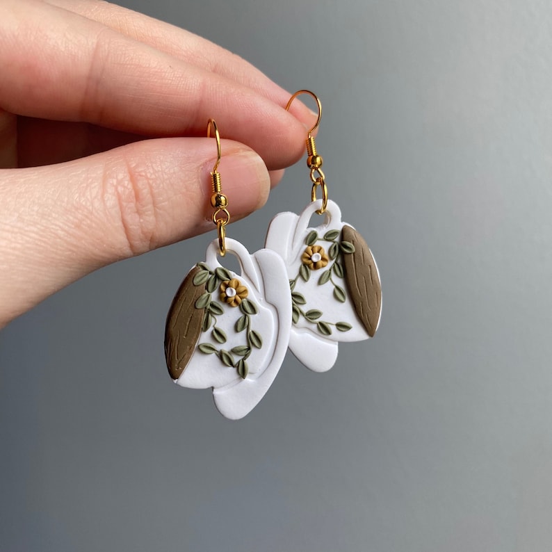Puede incluir: Un par de pendientes de cer&aacute;mica blanca con forma de taza de t&eacute; con detalles florales marrones y verdes. Los pendientes tienen ganchos dorados.